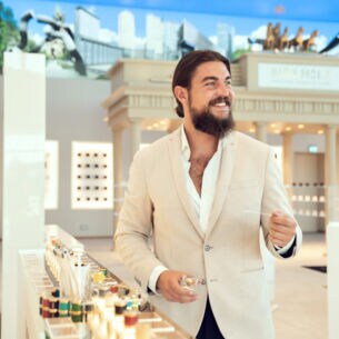 Ein Mann mit Vollbart in einem weißen Jackett steht im hellen Verkaufsraum einer Parfümmanufaktur und testet ein Parfüm mit Duft-Teststreifen