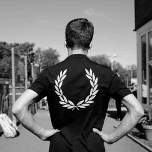Rückansicht eines Mannes in einem dunklen T-Shirt mit Lorbeerkranz-Logo-Druck auf dem Rücken auf einem Tennisplatz, Schwarzweißaufnahme