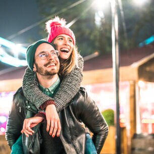 Ein Mann trägt eine Frau in weihnachtlicher Atmosphäre Huckepack, beide lächeln dabei