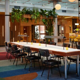 Langer Tisch mit Stühlen in einer Hotel-Lobby, im Hintergrund eine beleuchtete Bar