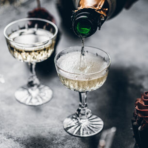 Zwei Gläser auf einem grauen Tisch werden mit Champagner gefüllt, daneben ist ein Cupcake mit Schokohaube zu sehen
