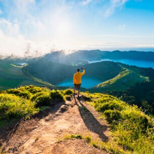Eine Person im gelben Oberteil steht auf einem Berg und blickt auf einen Vulkan auf den Azoren