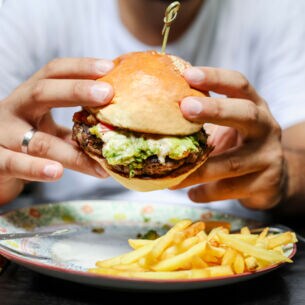 Eine Person hält einen Burger mit beiden Händen über einem Teller mit Pommes fest