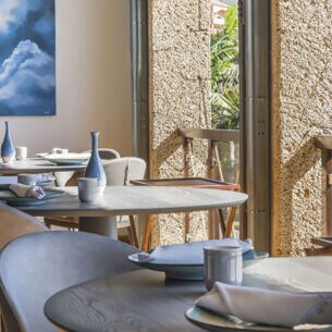 Innenraum eines luftigen, hellen Restaurants mit gedeckten Tischen an einer Fensterfront mit Blick auf Palmen