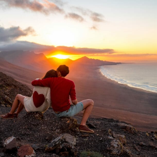 Zwei Personen sitzen mit dem Rücken zur Kamera auf einem Felshang und Blicken in den Sonnenuntergang über den Bergen, rechts im Bild das Meer