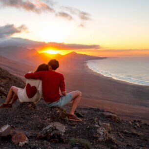 Zwei Personen sitzen mit dem Rücken zur Kamera auf einem Felshang und Blicken in den Sonnenuntergang über den Bergen, rechts im Bild das Meer