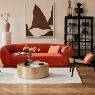 Ein modernes, helles Wohnzimmer mit einem geschwungenen, orangefarbenen Samtsofa vor einem runden, goldenen Couchtisch, daneben schwarze Möbelstücke als Akzent