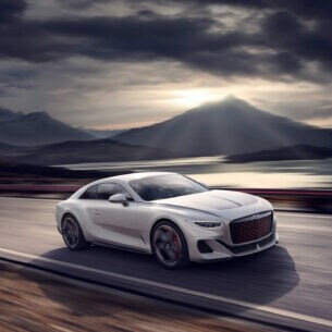 Weißes Auto von Bentley auf einer Straße, im Hintergrund Berge und Wolken
