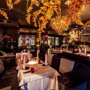 Ein Restaurant mit mehreren, weiß gedeckten Tischen, dunklen Sitzmöbeln und goldenen Leuchtern und Dekoblättern.