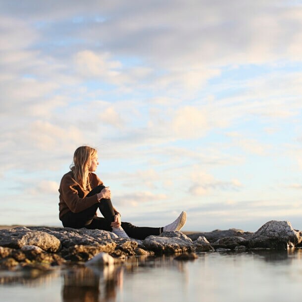 Junge Frau mit blonden langen Haaren sitzt auf Felsen und schaut übers Meer