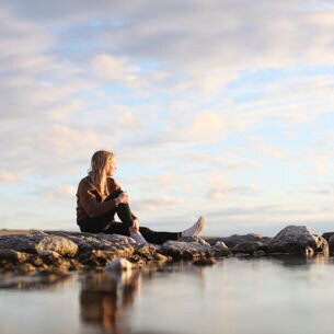 Junge Frau mit blonden langen Haaren sitzt auf Felsen und schaut übers Meer