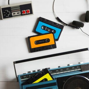 Auf einem Tisch liegen mehrere analoge Medien in Form von Kassetten, einer Polaroidkamera, einem Kassettenrekorder, einem NES-Gamepad und Joystick