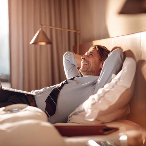 Ein Mann mit Hose, Hemd und Krawatte liegt lächelnd und mit hinter dem Kopf verschränkten Armen mit einem Laptop in einem Hotelbett.