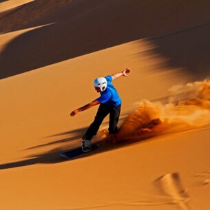 Ein Sandboarder mit Helm fährt eine rötliche Sanddüne hinunter