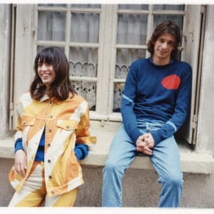 Ein Mann und eine Frau vor einem Fenster eines alten Gebäudes tragen farbige und lässige Outfits.
