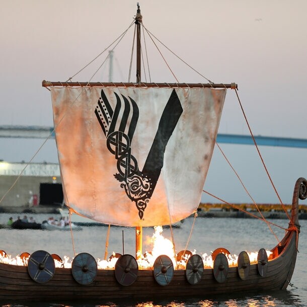 Aufnahme eines brennenden Wikingerschiffs auf dem Meer mit einem Segel, das das Logo der „Vikings“-Serie zeigt