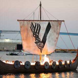 Aufnahme eines brennenden Wikingerschiffs auf dem Meer mit einem Segel, das das Logo der „Vikings“-Serie zeigt