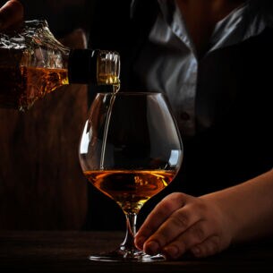 Ein Barkeeper hinter einer dunklen Holztheke gießt Cognac in ein bauchiges Glas