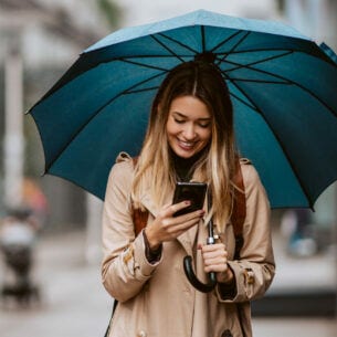 Eine junge Frau auf einer Straße schaut lächelnd auf ihr Smartphone unter einem Regenschirm