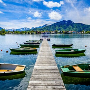 Ein Holzsteg mit kleinen Booten auf einem See, im Hintergrund Berge