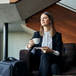 Eine junge Geschäftsfrau genießt die Wartezeit mit einer Tasse Kaffee auf einem Sessel in einer Lounge