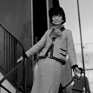Schwarzweißaufnahme von Coco Chanel in einem Tweed-Kostüm auf einer Treppe vor einer Spiegelwand