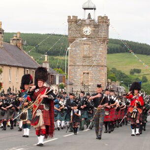 Diverse Menschen in traditionellen schottischen Outfits, die eine Straße entlanglaufen und Instrumente spielen.