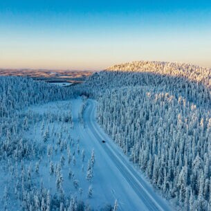 Luftaufnahme einer weiten Winterlandschaft mit schneebedeckten Wäldern, durch die ein einzelnes Auto über eine Straße durch den Schnee in der Abenddämmerung fährt