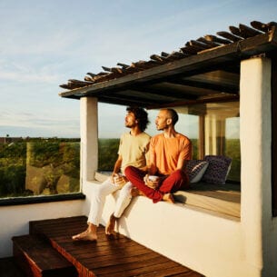 Zwei lässig gekleidete Männer sitzen auf einer Terrasse in der Abendsonne und schauen über eine tropische Landschaft aufs Meer