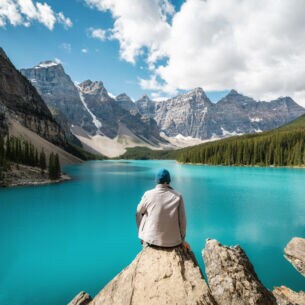 Eine Person sitzt auf einem Stein und schaut auf einen See, der gesäumt von bewaldeten Bergen ist