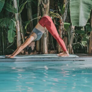 Eine junge Frau bei einer Yogaübung auf einer Matte an einem Pool, umgeben von Palmen