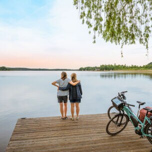 Rückansicht von zwei Frauen, die auf einem Steg vor einem See stehen und sich umarmen, daneben zwei E-Bikes