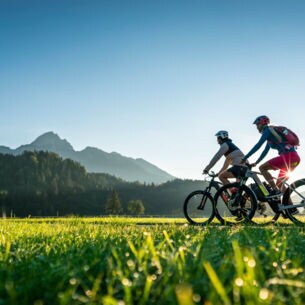 Zwei Personen fahren auf Mountainbikes durch eine Grasebene vor Bergpanorama im Sonnenschein