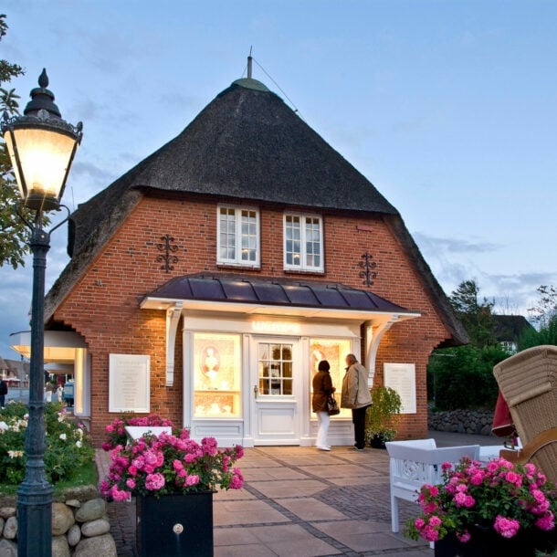 Zwei Personen stehen vor einem beleuchteten Schaufenster einer Luxusboutique in einem reetgedeckten Haus