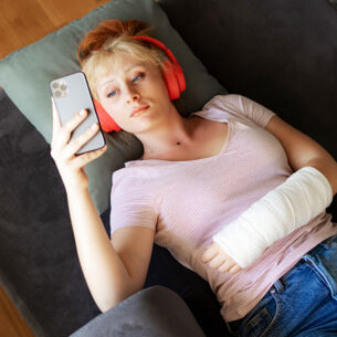 Aufsicht einer auf einem Sofa liegenden, jungen Frau, die mit Kopfhörern und einem eingegipsten Arm auf ihr Smartphone schaut.