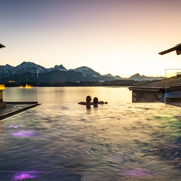 Zwei Personen in einem Infinity-Pool schauen über einen See in eine Berglandschaft bei Sonnenuntergang