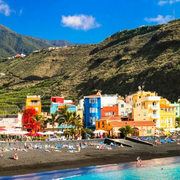 Schwarzer Sandstrand mit Badenden in einem Ort mit bunten Häusern vor Bergpanorama