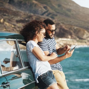 Zwei Personen stehen an ein Auto gelehnt an einer Meeresküste und schauen auf ein Smartphone