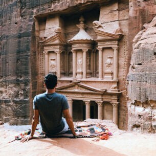 Rückansicht eines auf einem Felsvorsprung sitzenden Mannes vor dem Eingang eines monumentalen Grabtempels in der Felsenstadt Petra