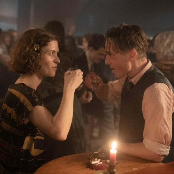 Ein belebter Nachtklub mit Tanzfläche, im Vordergrund ein Mann und eine Frau, die zusammen einen Schnaps trinken