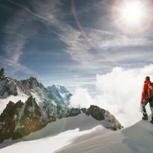 Zwei Personen gehen einen schneebedeckten Bergkamm entlang, im Hintergrund erhebt sich ein noch höheres Bergpanorama