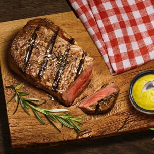 Aufsicht eines gebratenen, angeschnittenen Steaks auf einem Brett neben einer Schale mit Senf.