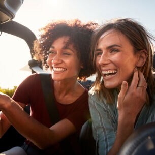 Nahaufnahme von zwei jungen, lachenden Frauen mit wehenden Haaren auf dem Rücksitz eines Cabriolets.
