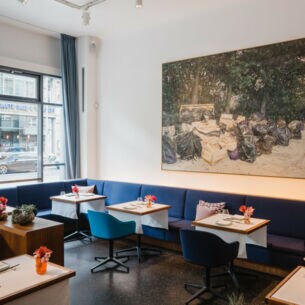 Innenraum eines hellen, modernen Restaurants mit blauen Sitzmöbeln an gedeckten Tischen und einem großen Gemälde an der Wand.