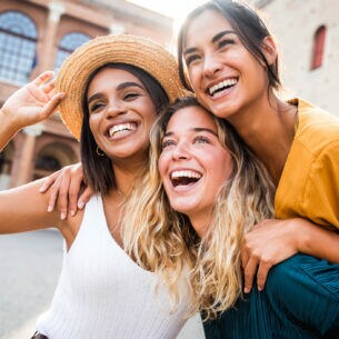 Drei lachende Freundinnen posierend umarmend für ein Foto auf einem öffentlichen Platz bei Sonnenschein.