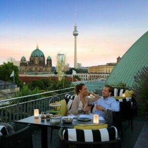 Ein junges Paar sitzt gut gelaunt an einem Tisch auf einer Dachterrasse, im Hintergrund Berliner Dom und Fernsehturm.