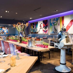 Restaurant mit einem langen, mit Pflanzen dekorierten Holztisch und einer Aufschnittmaschine mit Standfuß, umgeben von farbenfrohen Bildern an der Wand.