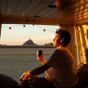 Ein junger Mann sitzt mit einem Kaffeebecher in der Hand im geöffneten Campervan im goldenen Sonnenlicht und blickt hinaus in die Natur.