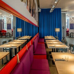 Modernes Restaurant mit hellen Holztischen und roten Sitzbänken an einer Spiegelwand.