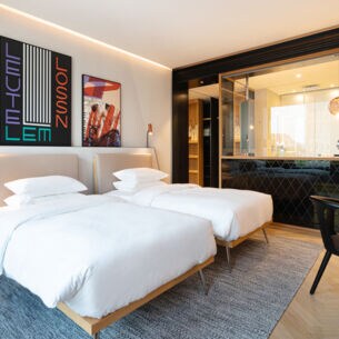 Elegantes Hotelzimmer im modernen Design mit zwei Betten und offenes Badezimmer hinter einer Glasscheibe.